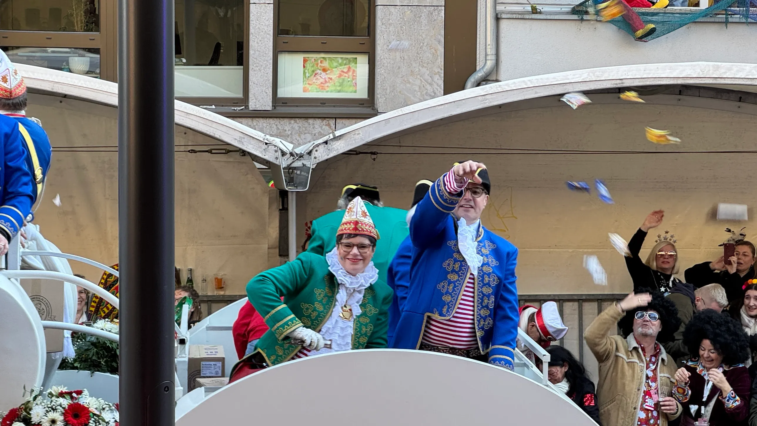 Zwei Karnevalisten in Grün und Blau werfen Kamelle vom Wagen, rechts jubelnde Zuschauer